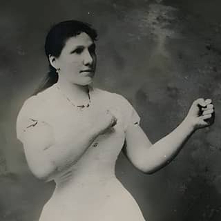 An old woman wearing a white dress holding a tennis racket and a boxing glove.