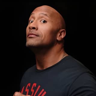 The rock is posing in a black t-shirt while a man holds a cell phone.