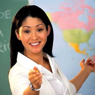Woman in white shirt smiling and pointing at camera in front of world map in classroom setting.