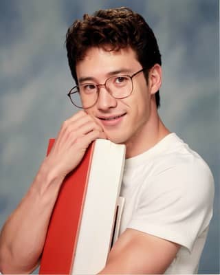 Un hombre con lentes está sonriendo mientras sostiene un libro, lo que sugiere que podría ser un estudiante en la Universidad de San Diego.