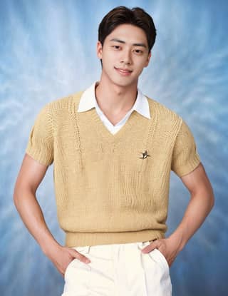 A man wearing a yellow sweater and white pants poses for a picture in a studio.