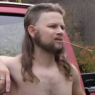 A person with a long hair and beard stands in front of a red truck while smoking a cigarette.