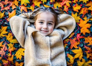 A little girl in a sweater laying on the ground surrounded by autumn leaves.
