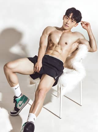 Modelo coreano de shorts pretos e sem camisa sentado em uma cadeira.