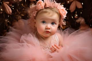 A baby girl in a pink tutu and flower crown sitting in front of a Christmas tree.