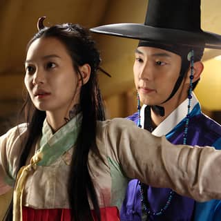 Two people dressed in traditional Korean clothing and hats, resembling the characters from the Korean movie 'The King.'