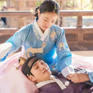 Woman in traditional Korean clothing, a blue and pink hanbok, laying on a bed.