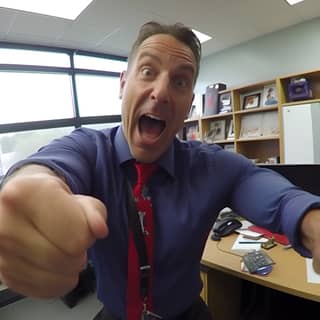 Realistica GoPro selfie di un preside delle scuole superiori sorridente che flette le braccia nella luce naturale del suo ufficio, indossando una camicia blu e una cravatta rossa.