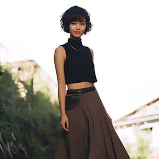Seorang wanita muda yang gembira dengan ciri etnik campuran, rambut pendek, dan memakai topi halter hitam, skirt panjang sebatas pergelangan kaki berwarna cokelat, dan bot tempur, berdiri di atas trotoar.