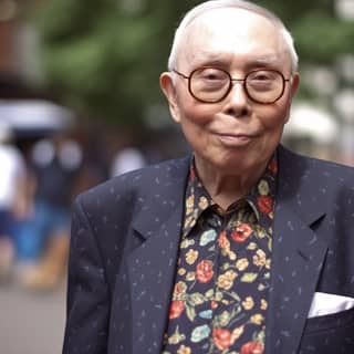 Un uomo anziano con gli occhiali e una camicia floreale si trova in strada, sembra curato e di buon umore.