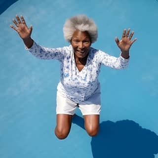 Una hermosa mujer negra de edad avanzada saltando en un trampolín al aire libre bajo un cielo azul.